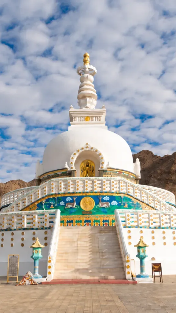 White Shanti Stupa in Leh surrounded by brown Himalayan mountains and cloudy blue sky